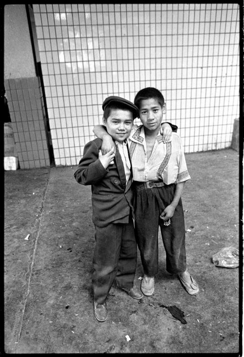 Photographie argentique en noir et blanc de deux jeunes garçons ouïgours, vendeurs de babioles, dans la gare routière de Kashgar en Chine.