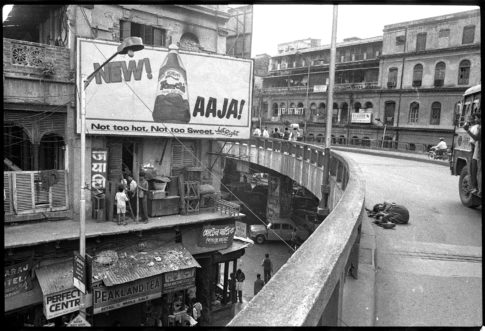 Photographie argentique en noir et blanc d'un quartier de Calcutta/Inde où s'entremêlent petits commerces animés, panneau publicitaire, axes routiers encombrés et homme gisant sur le bitume.