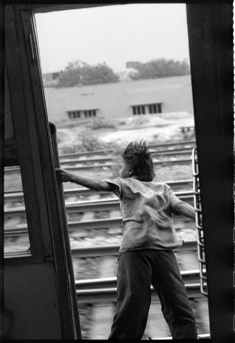 Photographie argentique en noir et blanc d'un garçon flottant dans le vide sur le marche pied d'un train lancé entre Bénarès et Delhi en Inde.